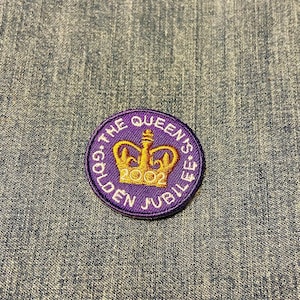 May include: A circular purple patch with gold text and a gold crown design. The text reads "THE QUEEN'S GOLDEN JUBILEE 2002". The patch is embroidered and has a stitched border, set against a denim background.