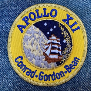 Peut inclure: Un écusson brodé jaune avec des lettres bleues qui lisent "Apollo XII Conrad Gordon Bean". L'écusson représente un navire blanc naviguant sur un fond bleu avec un croissant de lune et des étoiles.