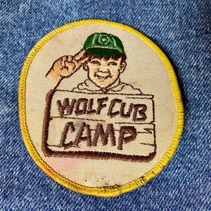 May include: A yellow embroidered patch with a cartoon boy wearing a green hat and holding a sign that says "Wolf Cub Camp".