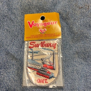 May include: A Voyageur patch with a cable car design and the word "Sudbury" in red script. The patch is in a clear plastic package, with "ONT." at the bottom. The patch is on a denim background.
