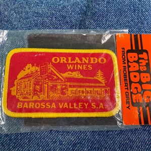May include: A red and yellow embroidered patch with the text "Orlando Wines Barossa Valley S.A." and a drawing of a building. The patch is packaged in a clear plastic bag with a red and black card that says "The Big Badge from Robert Grey".