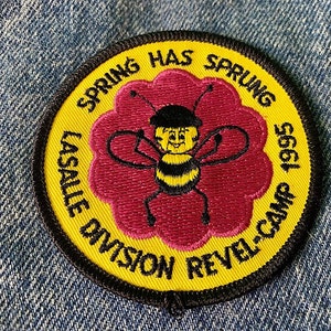 May include: Yellow and black embroidered patch with a cartoon bee in the center. The bee is wearing a black hat and has a smiling face. The text around the bee reads "Spring Has Sprung 1995 LaSalle Division Revel-Camp".