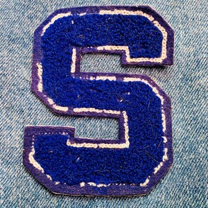 May include: A blue felt letter S with white stitching, on a denim background.