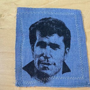 May include: A blue denim patch with a black and white image of a man's face. The patch is sewn with white thread around the edges.