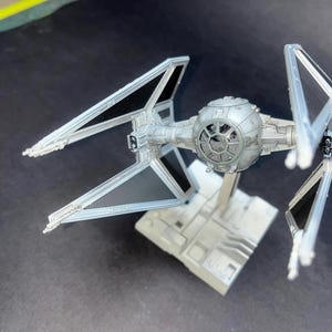 Könnte beinhalten: Detailliertes Modell eines TIE-Jägers, mit einem zentralen kugelförmigen Cockpit und zwei markanten Flügelpaneelen. Das Modell ist hauptsächlich grau mit schwarzen Akzenten und auf einem grauen Sockel montiert. Das Raumschiff ist ein Sammlerstück.