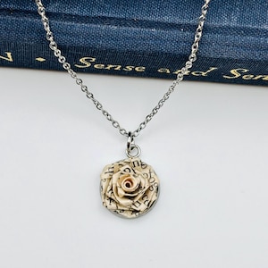 May include: Silver-toned necklace with a circular pendant featuring a paper rose. The rose is made from pages of a book, with visible text. The necklace is displayed against a backdrop of a book titled "Pride and Prejudice."