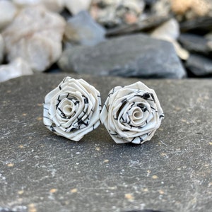 May include: A pair of white and black rose-shaped stud earrings. The earrings are made of a white material with black lines, creating a floral pattern. The earrings are displayed on a gray stone surface.