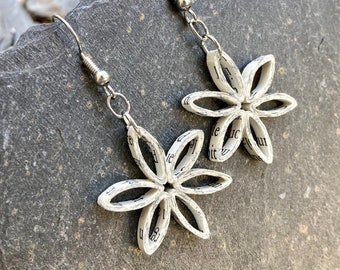 May include: A pair of handmade earrings featuring flower-shaped pendants. The pendants are crafted from rolled paper, displaying visible text, and are suspended from silver-colored hooks. The earrings rest on a gray stone surface.