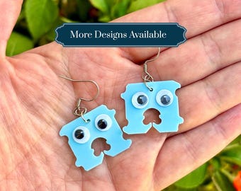 Op de afbeelding: Een paar lichtblauwe broodclip-oorbellen met bevestigde wiebeloogjes. Elke oorbel heeft een zilverkleurige metalen haak om op te hangen. De tekst "More Designs Available" staat op een donkerblauwe rechthoekige banner boven de oorbellen.