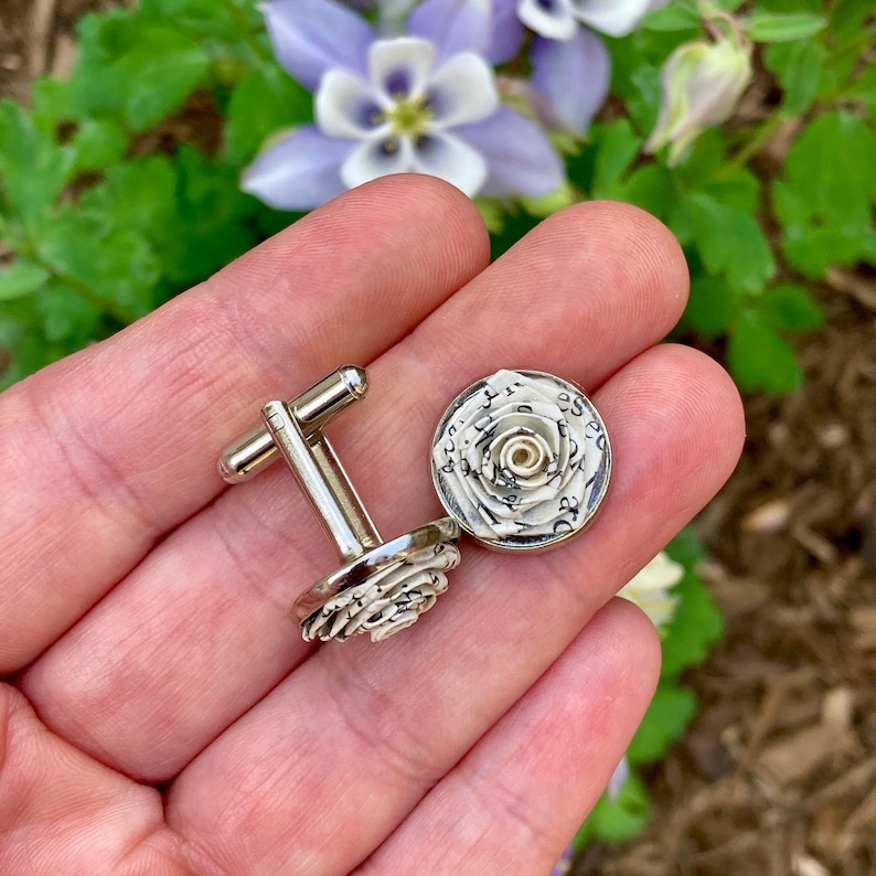 Handmade silver-tone cufflinks featuring miniature roses carefully crafted from recycled book pages, photographed against purple and white flowers.