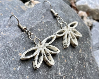 May include: A pair of handmade flower earrings. Each earring features a flower design crafted from rolled paper, with visible text. The earrings are suspended from silver-colored fish hook ear wires. The earrings are displayed on a gray stone surface.