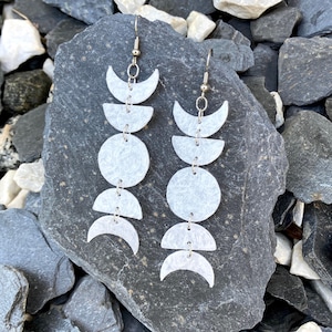 May include: White, layered moon phase earrings. Each earring features a crescent moon, a half-moon, and a full circle, all connected by silver-colored metal. The earrings are displayed on a gray stone surface.