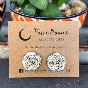 Op de afbeelding: Een paar oorknopjes van opgerold boekpapier in de vorm van rozetten, bevestigd op een rechthoekige bruine kaart met het opschrift Four Moons RECYCLED GIFTS, Handcrafted from book paper, en de website www.fourmoonsrecycled.com met iconen.