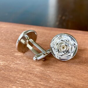 Op de afbeelding: Een paar zilverkleurige manchetknopen met een ronde voorkant voorzien van een driedimensionale roos gemaakt van wit papier bedrukt met zwarte tekst. Het metalen montuur heeft een standaard knevelsluiting.