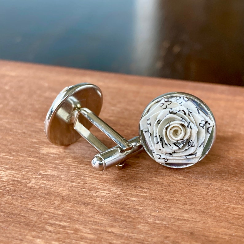 Op de afbeelding: Een paar zilverkleurige manchetknopen met een ronde voorkant voorzien van een driedimensionale roos gemaakt van wit papier bedrukt met zwarte tekst. Het metalen montuur heeft een standaard knevelsluiting.