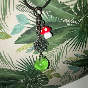 Puede incluir: Un llavero plateado con una cadena que sostiene un dije de hongo rojo y blanco, un dije de filigrana plateada y un dije de rana verde. El fondo presenta un patrón de hojas tropicales.