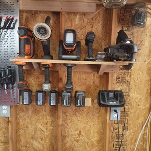 May include: A collection of orange and black power tools, including a drill, grinder, and work light, displayed on a wooden shelf. Several battery packs and a charger are also visible, all against a wood-paneled wall.