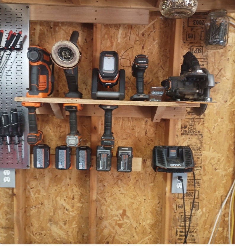 May include: A collection of orange and black power tools, including a drill, grinder, and work light, displayed on a wooden shelf. Several battery packs and a charger are also visible, all against a wood-paneled wall.
