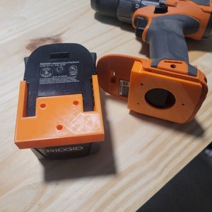 May include: An orange 3D-printed battery holder and a drill. The holder is designed to fit a Ridgid battery. The drill is black and orange. The battery holder has three holes for mounting.