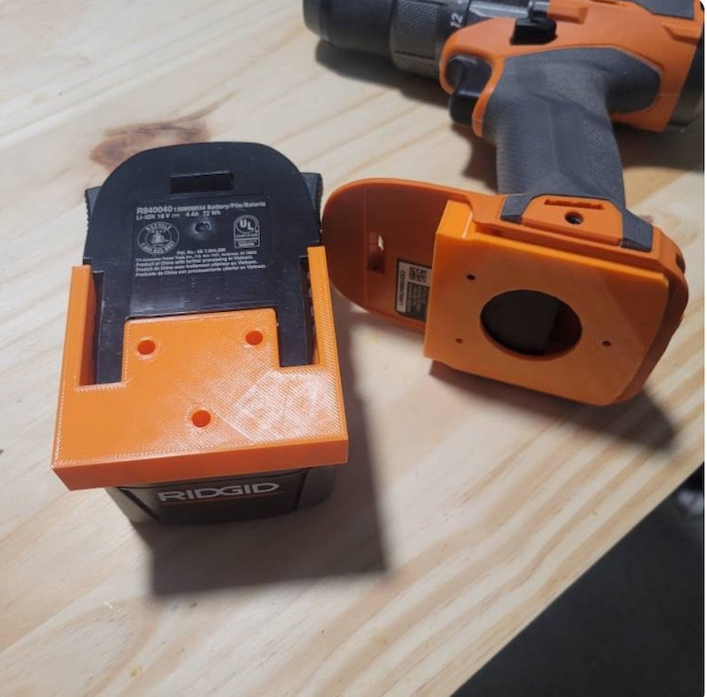 May include: An orange 3D-printed battery holder and a drill. The holder is designed to fit a Ridgid battery. The drill is black and orange. The battery holder has three holes for mounting.
