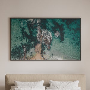 May include: An aerial photograph of a rocky coastline with turquoise water. The image is framed in a black frame and hangs on a white wall.