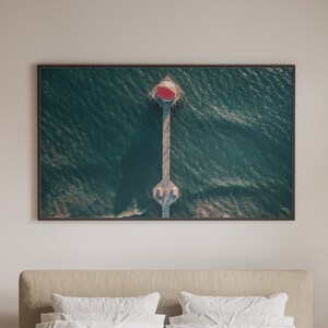 May include: A framed aerial photograph of a pier extending into the ocean. The pier has a red and white structure at the end. The water is a deep teal color, and the image is framed in a dark brown border. The photograph is displayed above a bed.