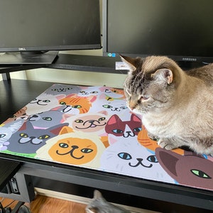 Puede incluir: Dos gatos tabby grises en un escritorio negro con una alfombrilla de ratón con temática de gatos. La alfombrilla de ratón presenta un patrón colorido de caras de gatos de dibujos animados. El escritorio tiene dos monitores de computadora en el fondo.