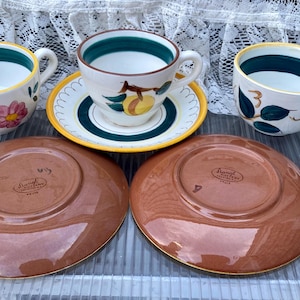 May include: A set of vintage teacups and saucers. The cups feature floral and fruit designs with green and yellow accents. The saucers are brown with a gold rim. The set is perfect for serving tea.