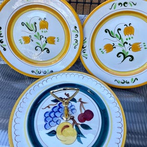 May include: Three ceramic plates with hand-painted floral designs. The plates have a white base with yellow and green accents. One plate is a serving platter with a gold handle and a painted fruit design. The plates are likely for decorative or serving purposes.