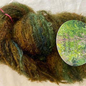 May include: A skein of hand-dyed yarn in shades of green, brown, and blue. The yarn has a textured, fluffy appearance. A round tag with the text "Solidago Treasures" is attached with pink and green string.