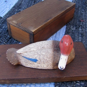 May include: A wooden duck carving with a red head, white beak, and gray body, resting on a dark brown wooden base. A rectangular wooden box sits in the background. The carving has a blue accent on the wing.