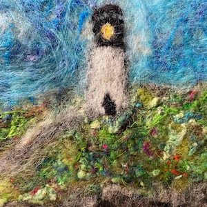 May include: A felted art piece depicting a lighthouse on a grassy hill under a blue sky. The lighthouse is white with a black top and a yellow light. The grass is green and colorful, and the sky is a mix of blue and white.