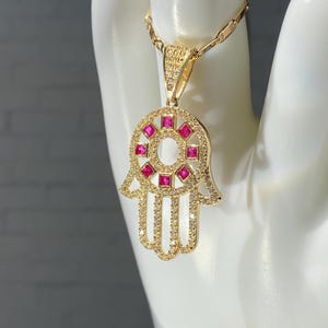 May include: A gold-colored Hamsa hand pendant necklace. The pendant features a central design of pink square gemstones surrounded by smaller clear stones. The necklace is displayed on a white surface.