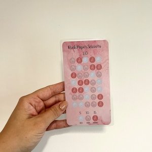 May include: A pink card with the text "Rock Paper Scissors 2.0" and hand symbols representing rock, paper, and scissors. The card has a marble-like background and is held by a hand.