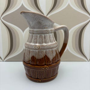 May include: A ceramic jug with a barrel-shaped design. The jug has a brown base with a wood grain texture, a light brown middle section, and a speckled white top. It has a curved handle and a spout for pouring liquids.
