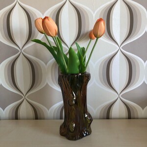 Könnte beinhalten: Eine braune Keramikvase in Form eines Baumstamms mit rauer Textur. Die Vase ist mit orangefarbenen Tulpen gefüllt.