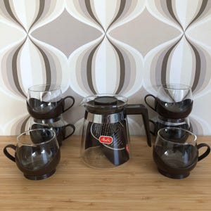 Könnte beinhalten: Ein Set aus sechs braunen und klaren Glaskaffeebechern und einer Glaskanne mit braunem Deckel und Griff. Die Kaffeekanne hat ein Etikett mit der Aufschrift "Melitta". Die Becher und die Kanne stehen auf einer Holzoberfläche.