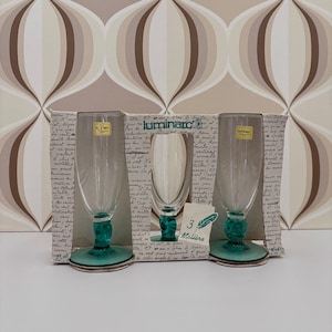 Op de afbeelding: Drie Luminarc champagneglazen met heldere glazen kommen en teal groene voetjes en stelen. De glazen worden weergegeven in hun originele verpakking, met het woord "Luminarc" zichtbaar. De verpakking heeft een vintage design.