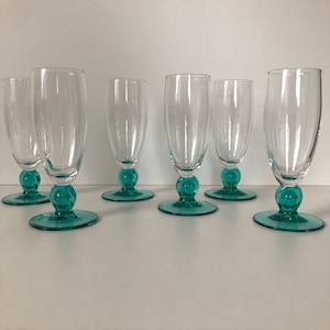 Op de afbeelding: Zes heldere glazen champagneflutes met tealblauwe voetjes. De flutes zijn gerangschikt in twee rijen van drie.