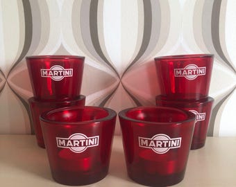 Puede incluir: Un conjunto de portavelas de cristal rojo con el logotipo blanco "MARTINI". Los portavelas están apilados. La imagen muestra una colección de estos portavelas sobre una superficie de color claro.