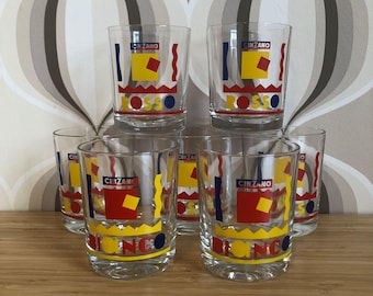 Puede incluir: Juego de seis vasos transparentes con el logotipo de Cinzano en rojo, amarillo y azul. Los vasos tienen un diseño retro con las palabras "ROSSO" y "BIANCO" impresas. Los vasos están colocados sobre una superficie de madera.