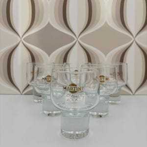 Op de afbeelding: Set van zes heldere Martini-glazen met een matte basis. Elk glas heeft het Martini-logo in goud en de tekst "ON THE ROCKS". De glazen staan op een wit oppervlak.