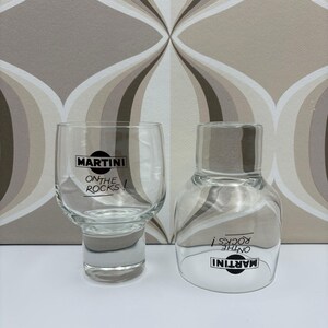Op de afbeelding: Twee heldere Martini-glazen, één een tumbler en de andere een afgerond glas met korte steel, beide met het Martini-logo en "ON THE ROCKS!" in zwart gedrukt. De achtergrond heeft een retro patroon in bruin- en wittinten.