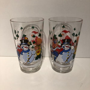 May include: Two clear glass tumblers featuring a winter scene design. The design depicts a snowman, Santa Claus, and other winter characters in a snowy landscape. The glasses are ideal for festive occasions.
