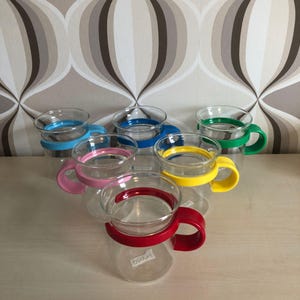 Op de afbeelding: Een verzameling heldere glazen mokken met gekleurde handvatten en banden in blauw, roze, geel, groen en rood. De mokken hebben een afgeronde vorm en zijn ontworpen voor warme dranken. De mokken zijn gerangschikt op een lichtgekleurd oppervlak.