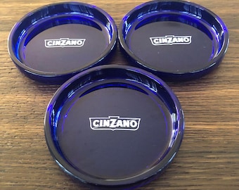 Puede incluir: Tres posavasos redondos azul marino con el logotipo "CINZANO" en blanco. Los posavasos tienen un acabado brillante y están dispuestos sobre una superficie de madera. Accesorio de bar.