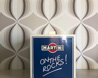 Puede incluir: Un letrero cuadrado blanco con fondo azul y el texto "MARTINI ON THE ROCKS!" en blanco. El logotipo de Martini es un círculo rojo con la palabra "MARTINI" en blanco.