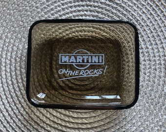 Puede incluir: Un cenicero de vidrio marrón con un borde negro. El cenicero tiene el logotipo de Martini y el texto "ON THE ROCKS!" impreso en blanco.