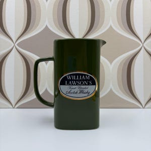 Pode incluir: Jarro de cerâmica verde escuro com uma pega e um bico. O jarro tem um rótulo oval com o texto "WILLIAM LAWSON'S Finest Blended Scotch Whisky" em letras brancas e douradas. Sobre uma superfície branca.