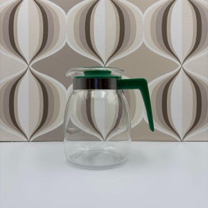Puede incluir: Una jarra de café de vidrio transparente con asa y tapa verdes. La jarra tiene una banda plateada alrededor del centro. El asa y la tapa son de color verde brillante. La jarra está sobre una superficie blanca.